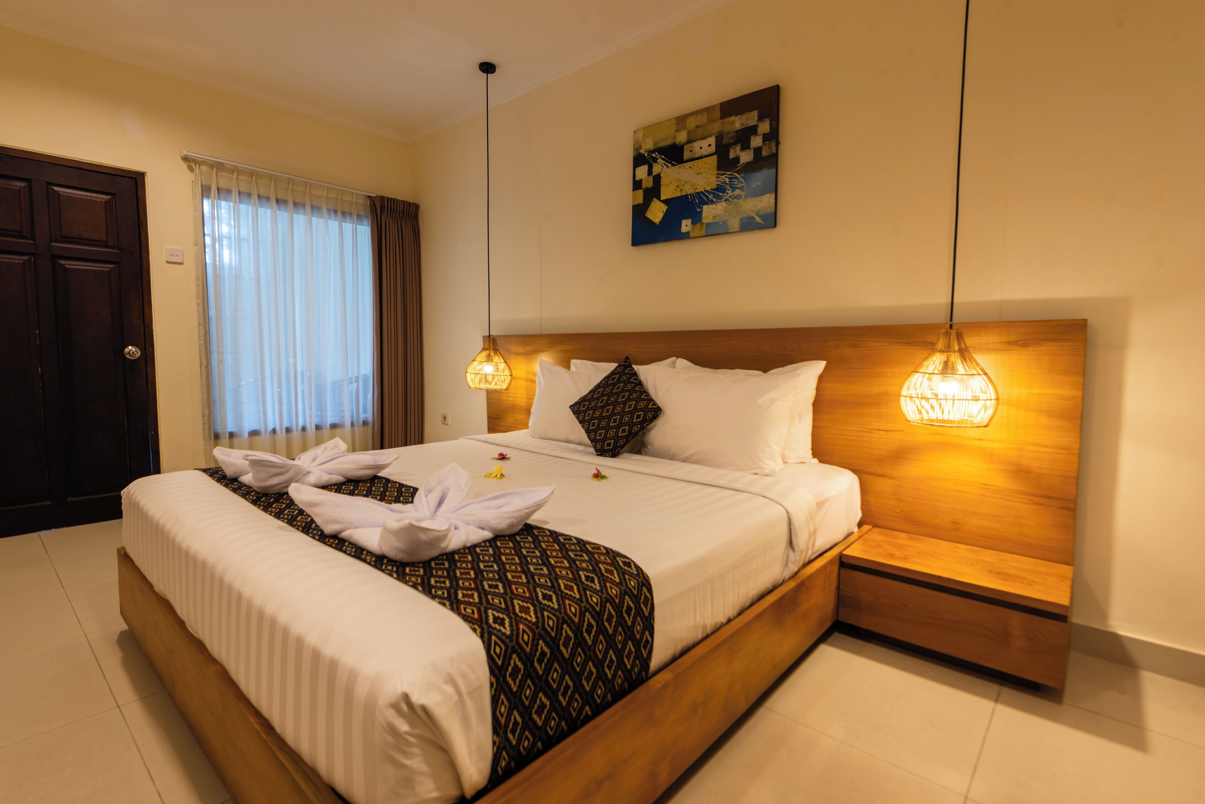 A hotel room with a large bed, wooden headboard, two bedside lamps, a painting on the wall, and a door.