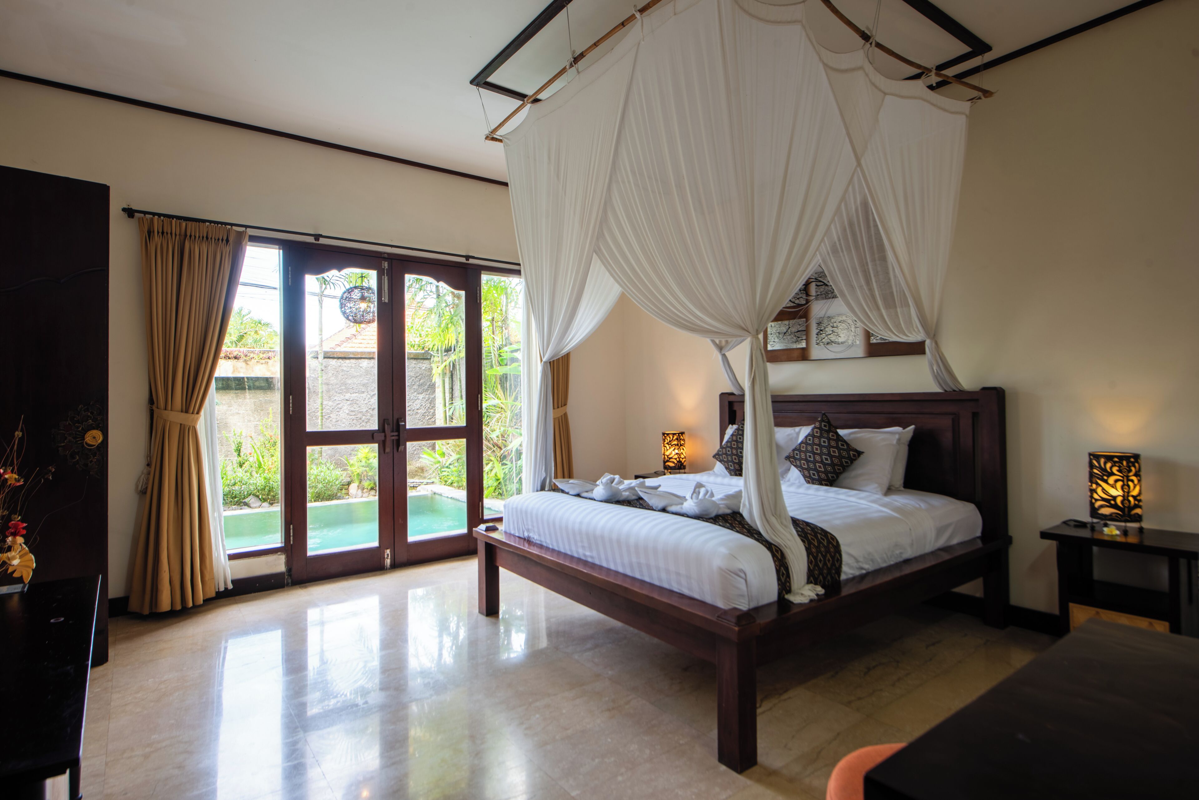 A bedroom with a canopy bed, a large sliding glass door leading to a pool, and a small table with a lamp.
