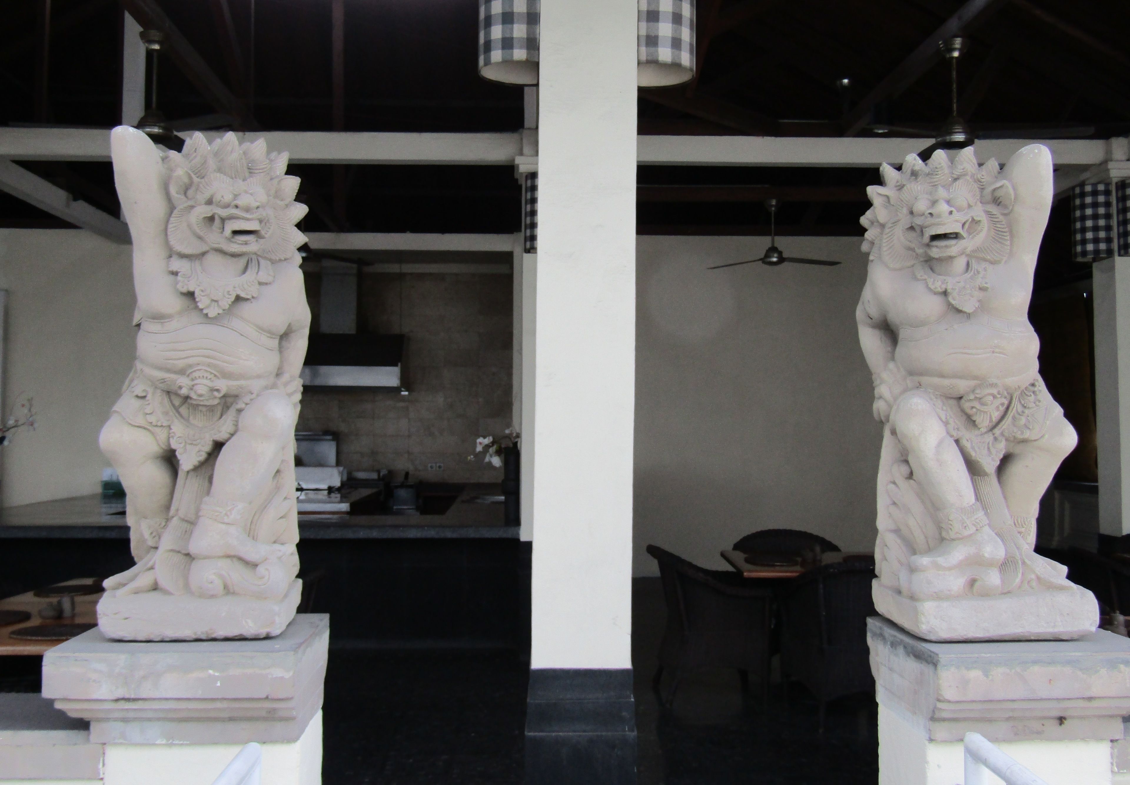 Deux statues de pierre blanche se trouvent dans une pièce au décor moderne, comprenant un ventilateur de plafond et, au fond, un coin cuisine.