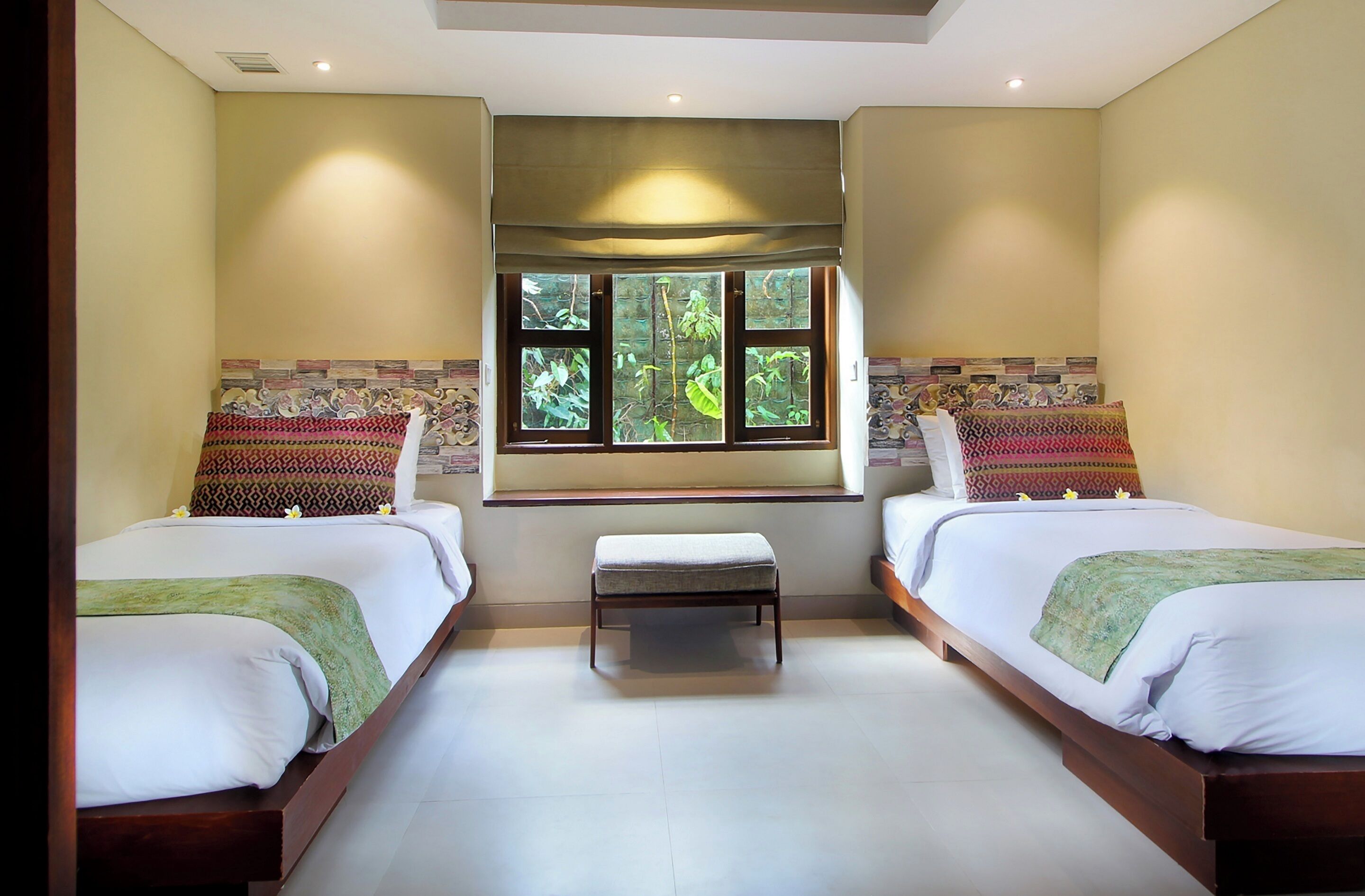 A hotel room with two beds, a bench, a window with a view of greenery, and a patterned wall.