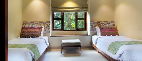 A hotel room with two beds, a bench, a window with a view of greenery, and a patterned wall.