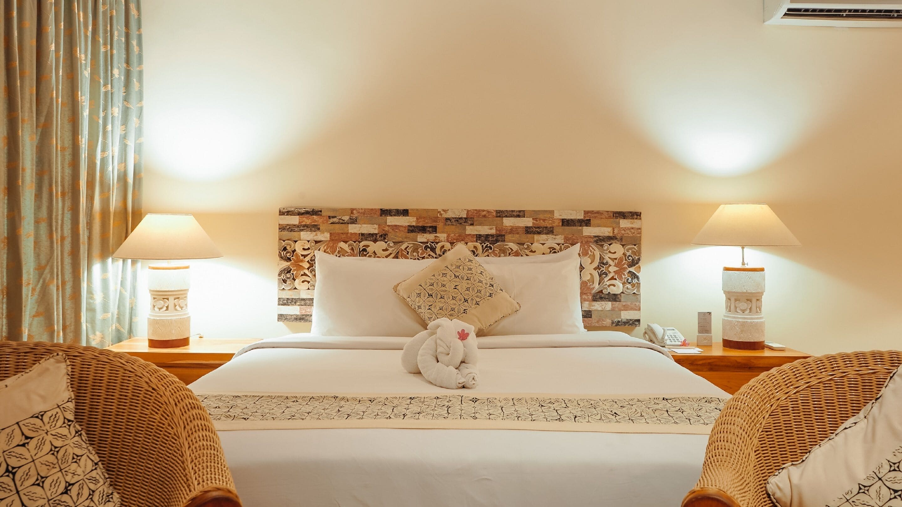 A hotel room with a neatly made bed, two bedside lamps, a decorative headboard, and a woven chair.
