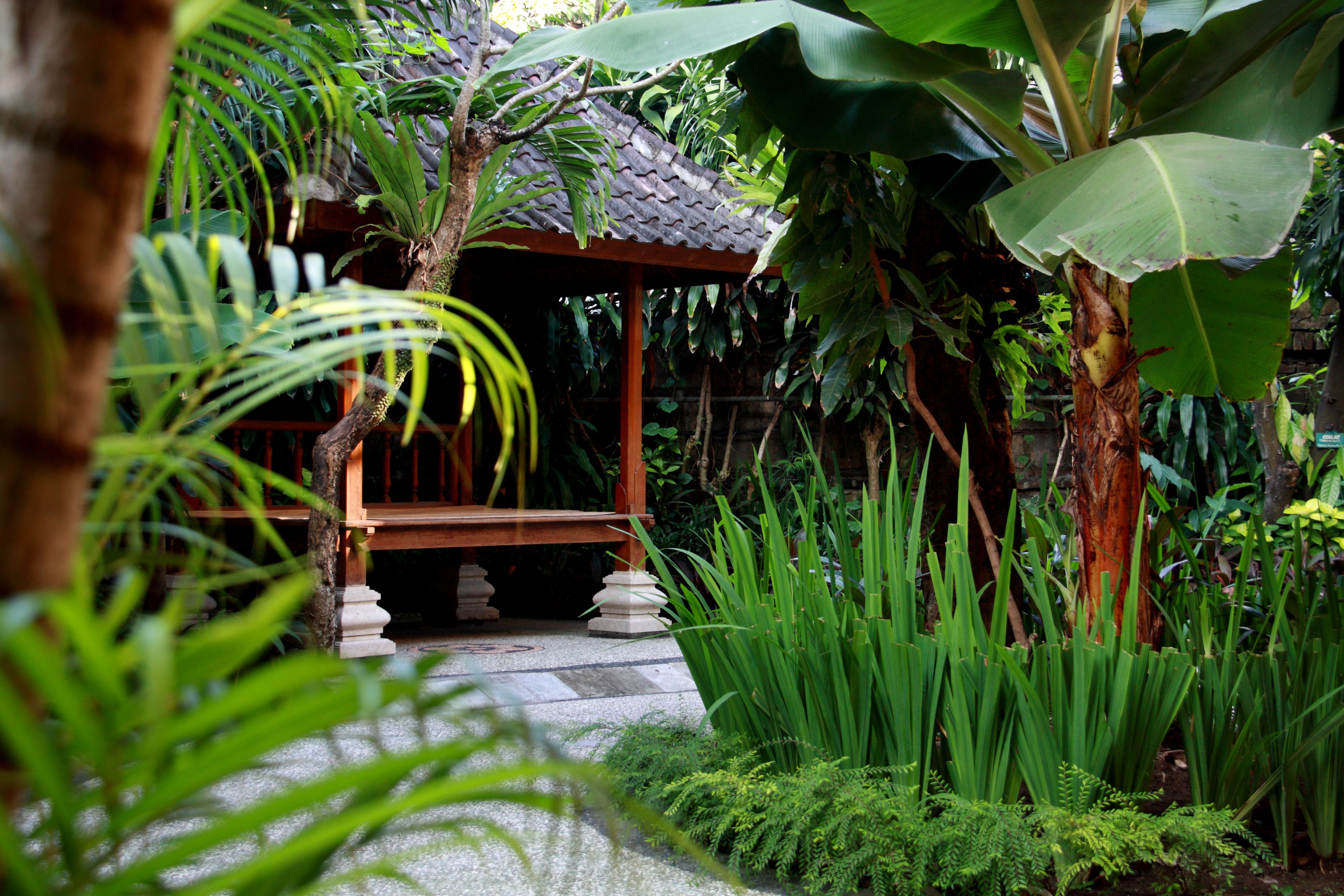 A wooden gazebo with a tiled roof surrounded by lush greenery and a stone pathway.