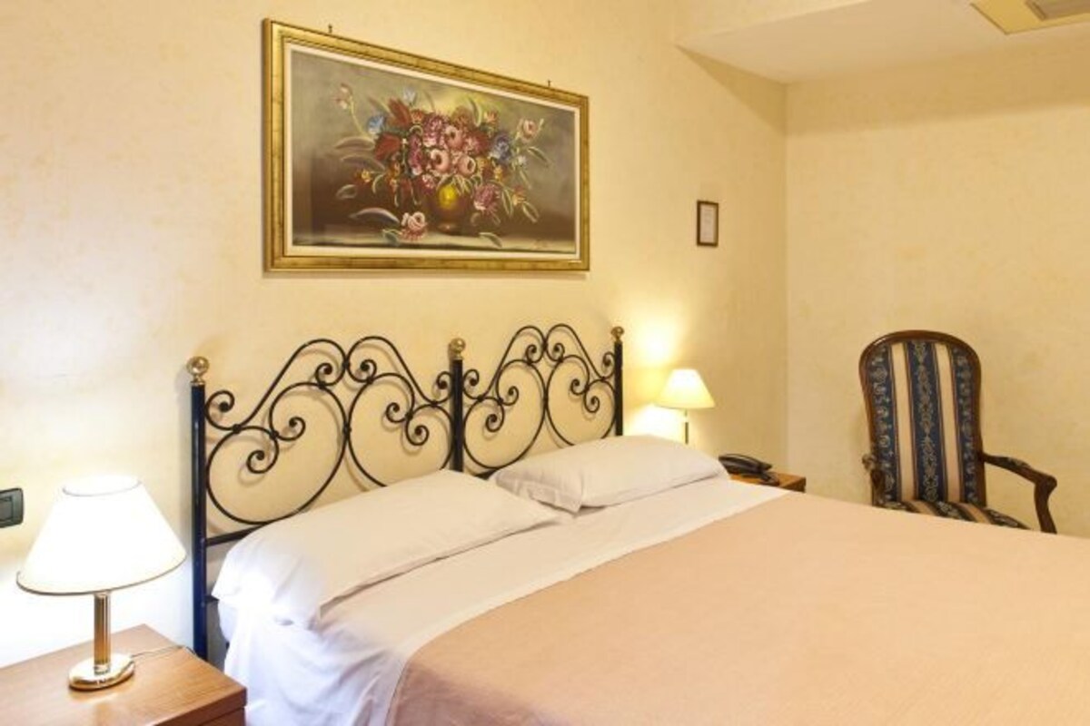 A neatly made bed with a headboard, a bedside lamp, a chair, and a framed floral painting on the wall.