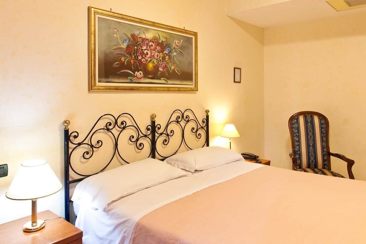 A neatly made bed with a metal headboard, a bedside lamp, a chair, and a framed painting on the wall.