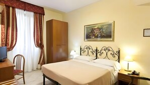 Une chambre d’hôtel comprenant un lit, un bureau, une chaise, une armoire, un tableau au mur et une table de chevet avec une lampe.