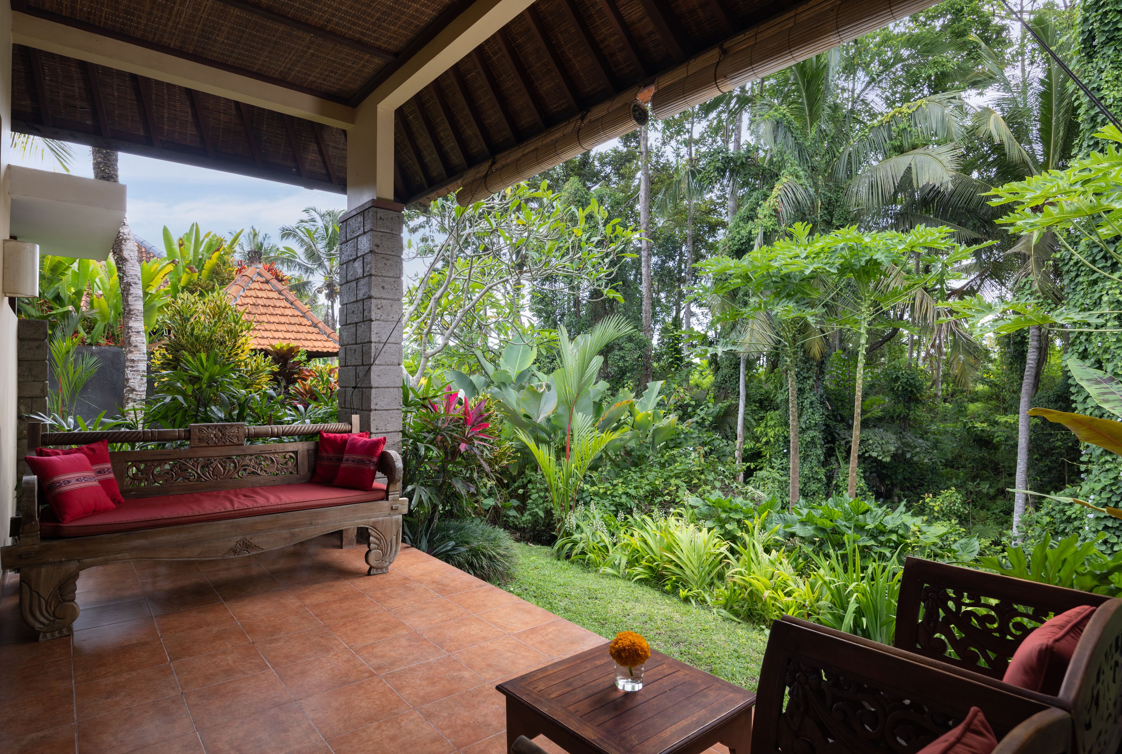 Un área de estar al aire libre techada con un banco y una mesa, rodeada de exuberante vegetación tropical y vista a una casa tradicional.