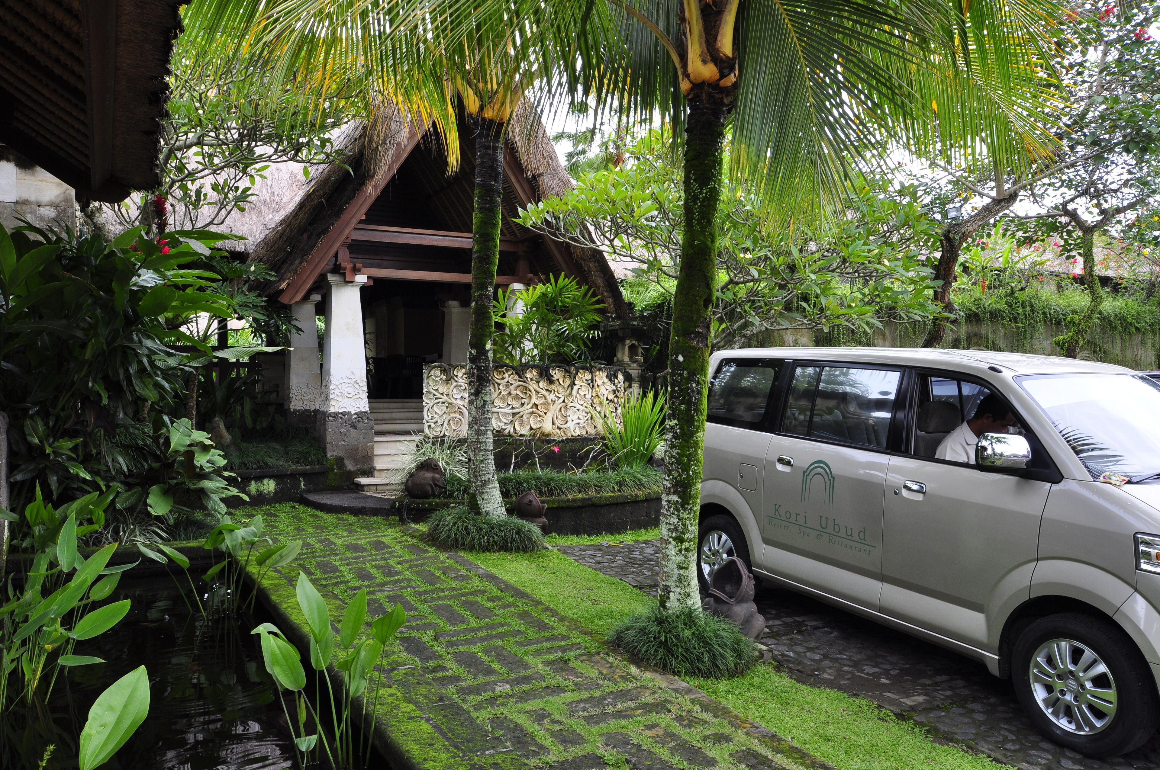 Un vehículo estacionado en un jardín exuberante con una estructura de techo de paja, rodeado de plantas tropicales y un pequeño elemento acuático.