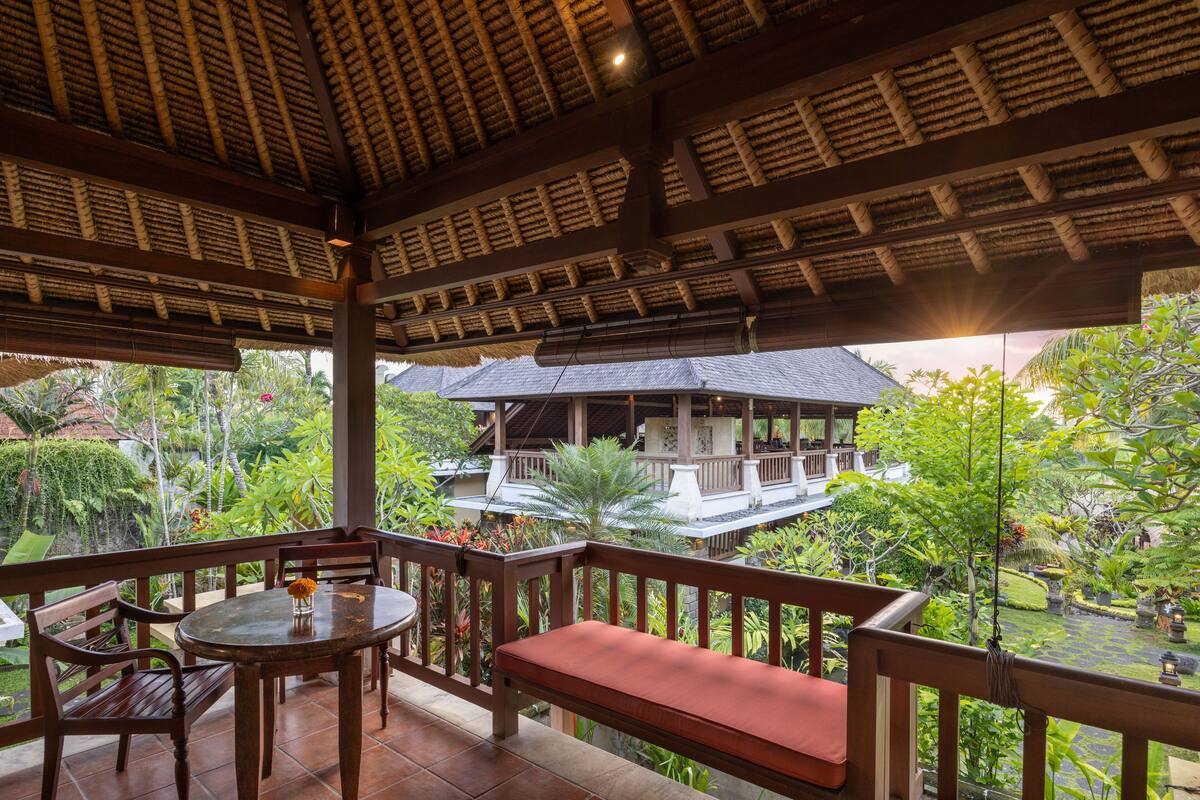 A covered outdoor seating area with a table, chairs, and a bench, overlooking a garden and a building with a thatched roof.