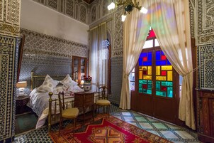 Quarto com cama, cadeira de madeira, mesa com um vaso e janela com vitrais.