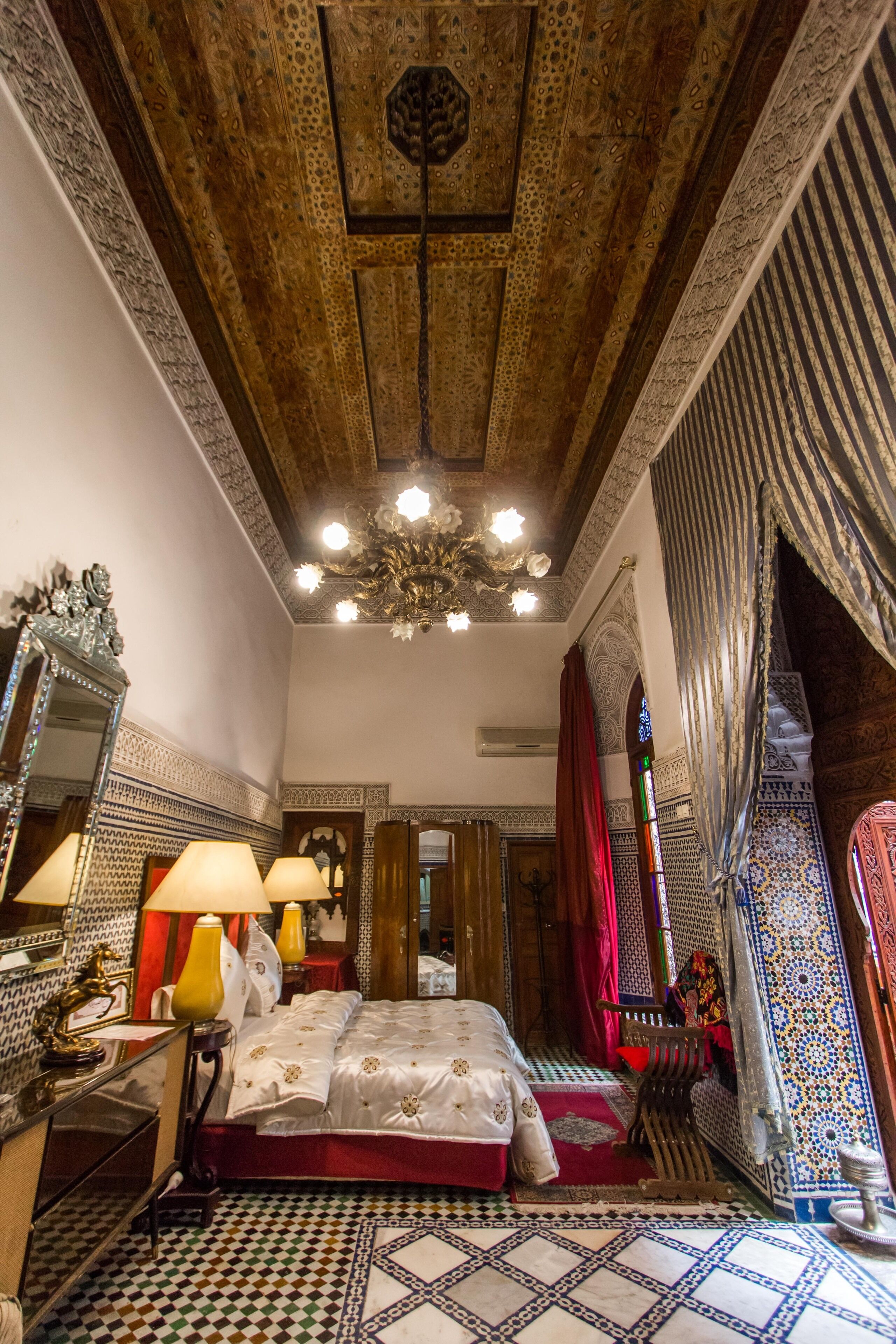 A traditional room with a patterned floor, a bed with a red bedspread, a chandelier, and a mirror with a decorative frame.