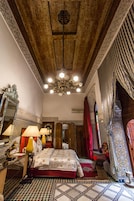 Una habitación tradicional con piso estampado, cama con colcha roja, candelabro y espejo con marco decorativo.