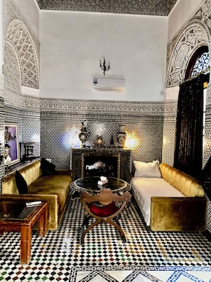 Um quarto com piso xadrez, um sofá, uma mesa com almofada vermelha e uma lareira.