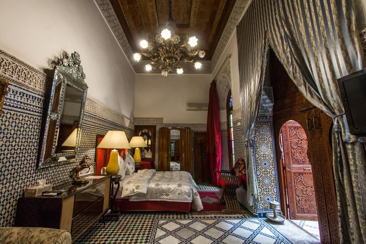 A traditional room with a bed, two lamps, a mirror, and patterned walls.