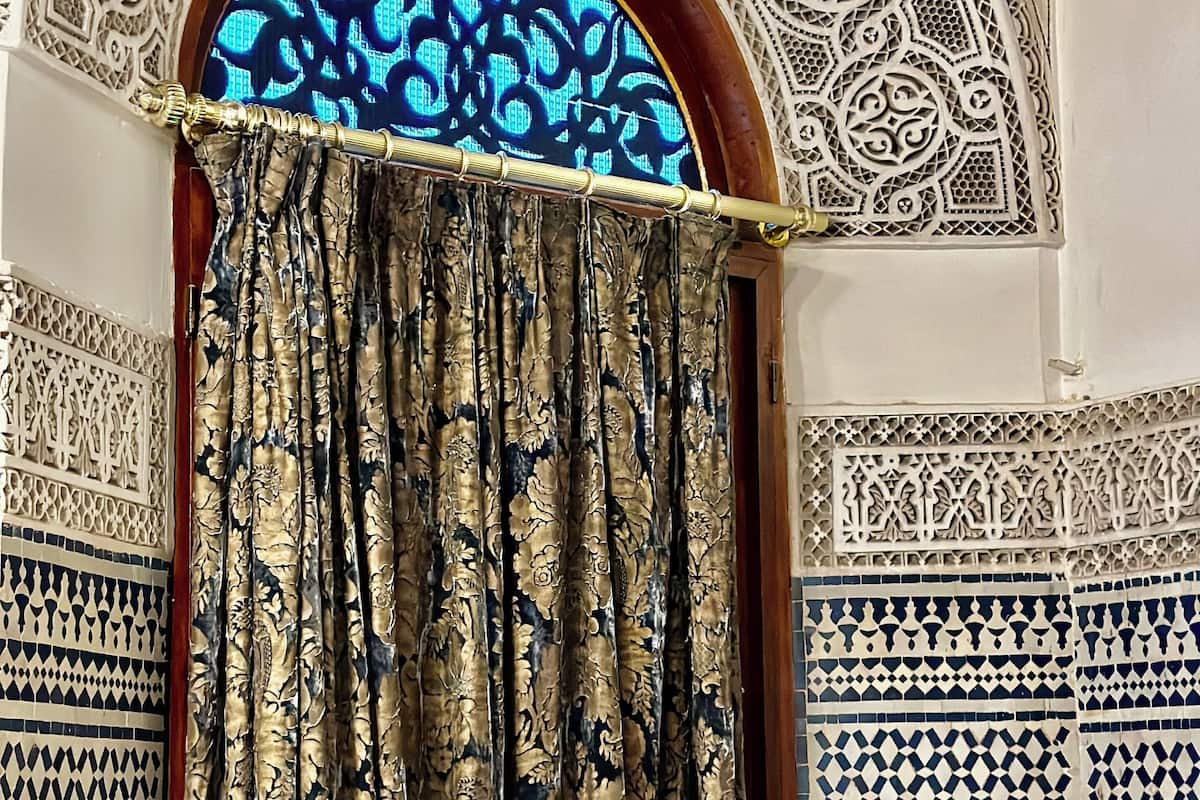 A room with intricate geometric and floral wall patterns, a wooden-framed window with a blue patterned view, and a curtain with a floral design.