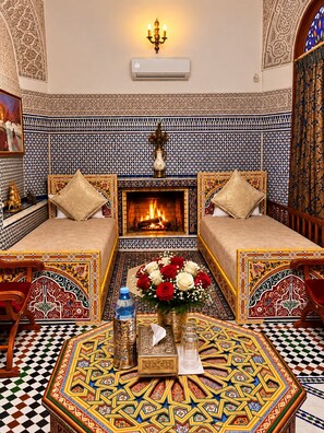 A traditional Moroccan room with a patterned tile wall, a fireplace, two sofas, a coffee table, and a decorative vase. - Riad Damia Suite &Spa (Fes)