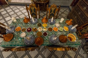 Uma mesa de jantar tradicional marroquina com vários pratos e utensílios, rodeada por cadeiras ornamentadas e um chão com padrão.