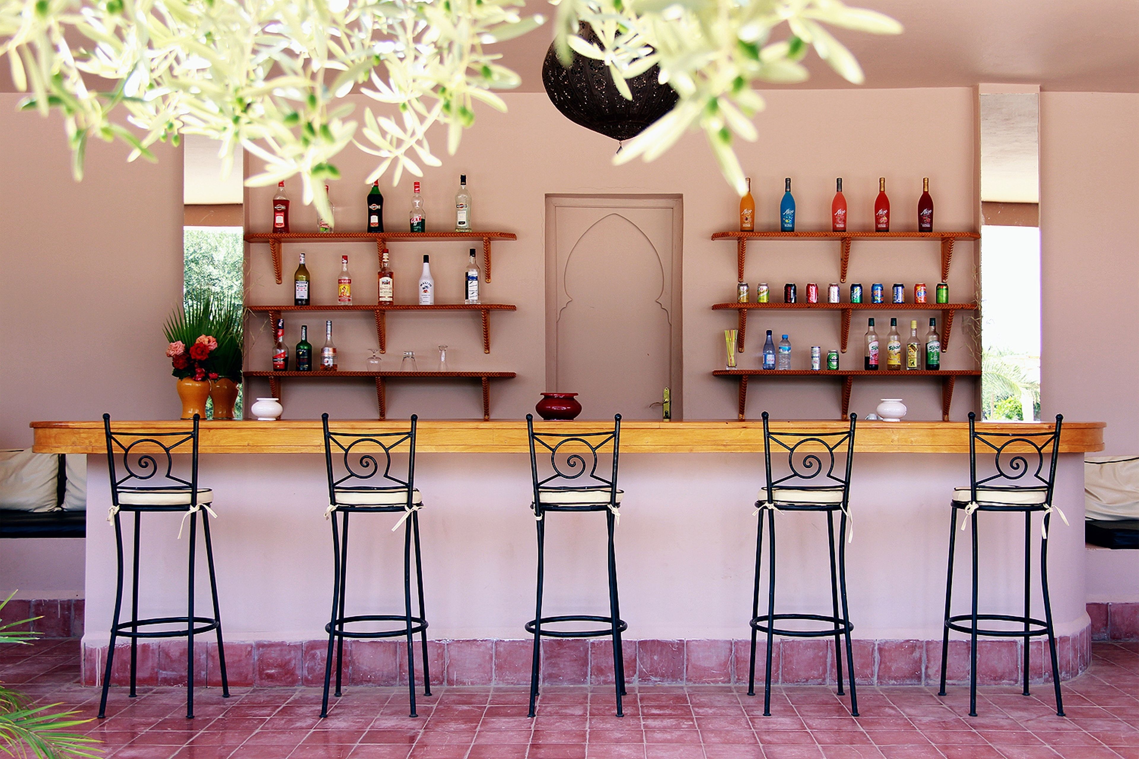 A bar area with a wooden counter, four metal barstools, and shelves stocked with various bottles and glasses.