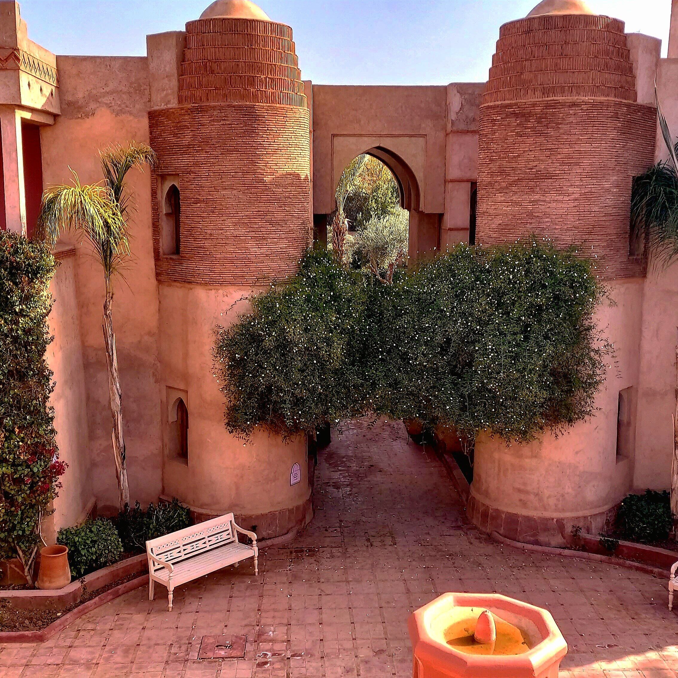 Un patio con un arco central, rodeado de edificios de ladrillo con techos abovedados, un banco y plantas en macetas.