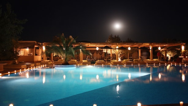 Ein Schwimmbecken bei Nacht mit Mond am Himmel, umgeben von Liegebereichen mit Sonnenschirmen und Kerzen.