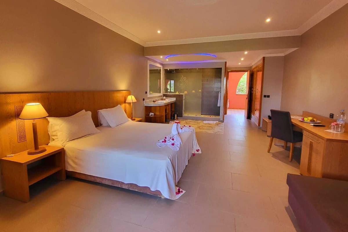 Une chambre d’hôtel avec un grand lit, des tables de chevet avec des lampes, un bureau avec une chaise, un espace douche et une vue sur l’extérieur.