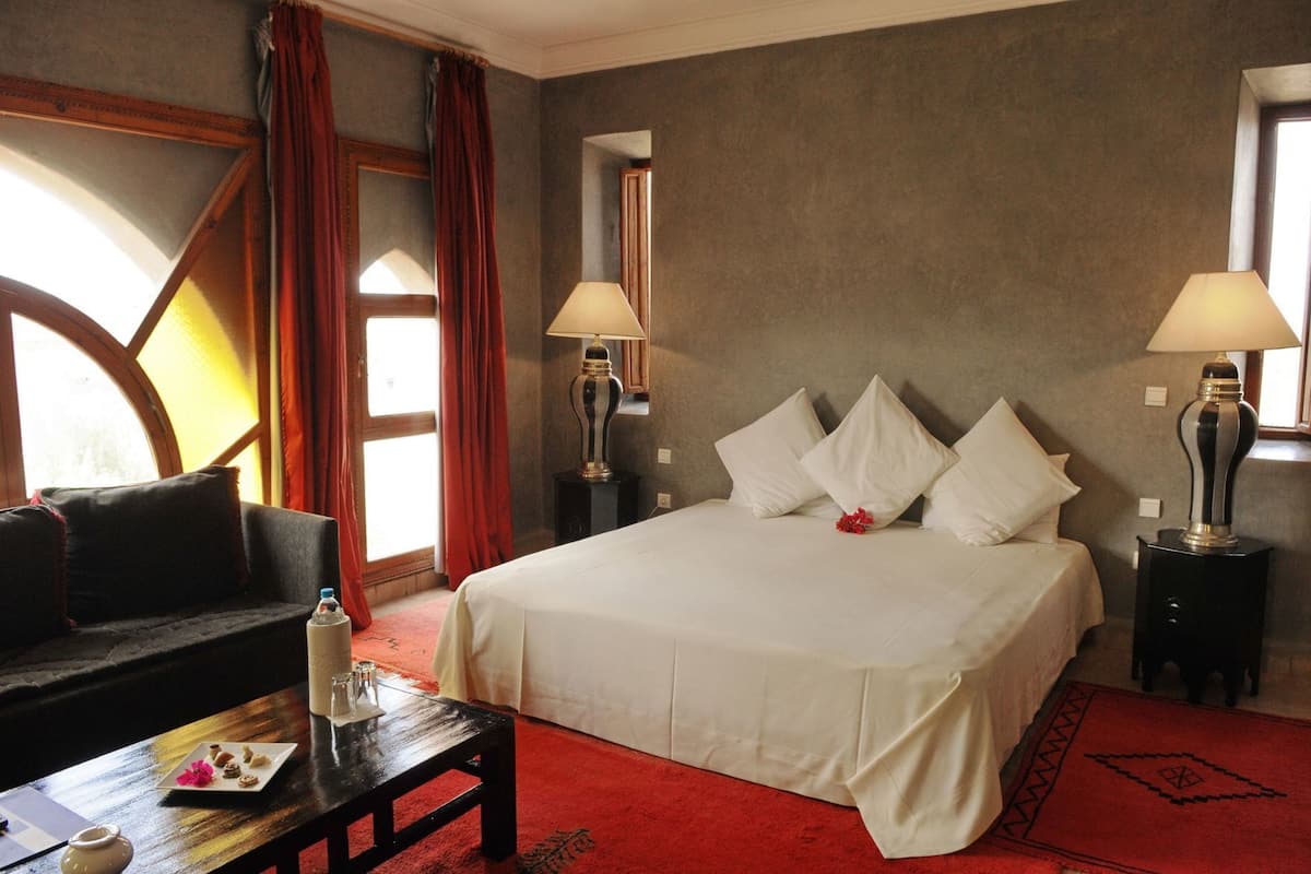 Une chambre d’hôtel avec un lit, deux lampes de chevet, un canapé, une petite table sur laquelle se trouvent un vase et une assiette de pâtisseries, et une moquette rouge.