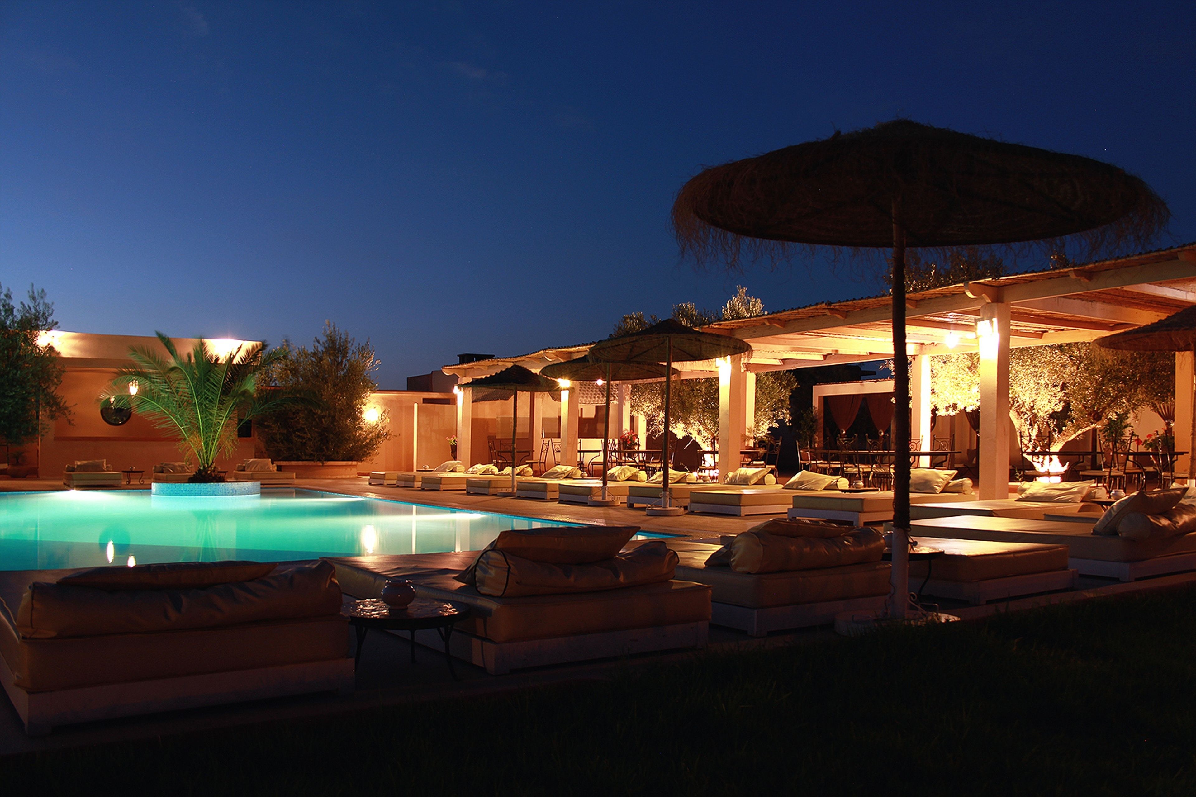 Un área junto a la piscina con sillones de descanso y sombrillas, rodeada por un porche cubierto.
