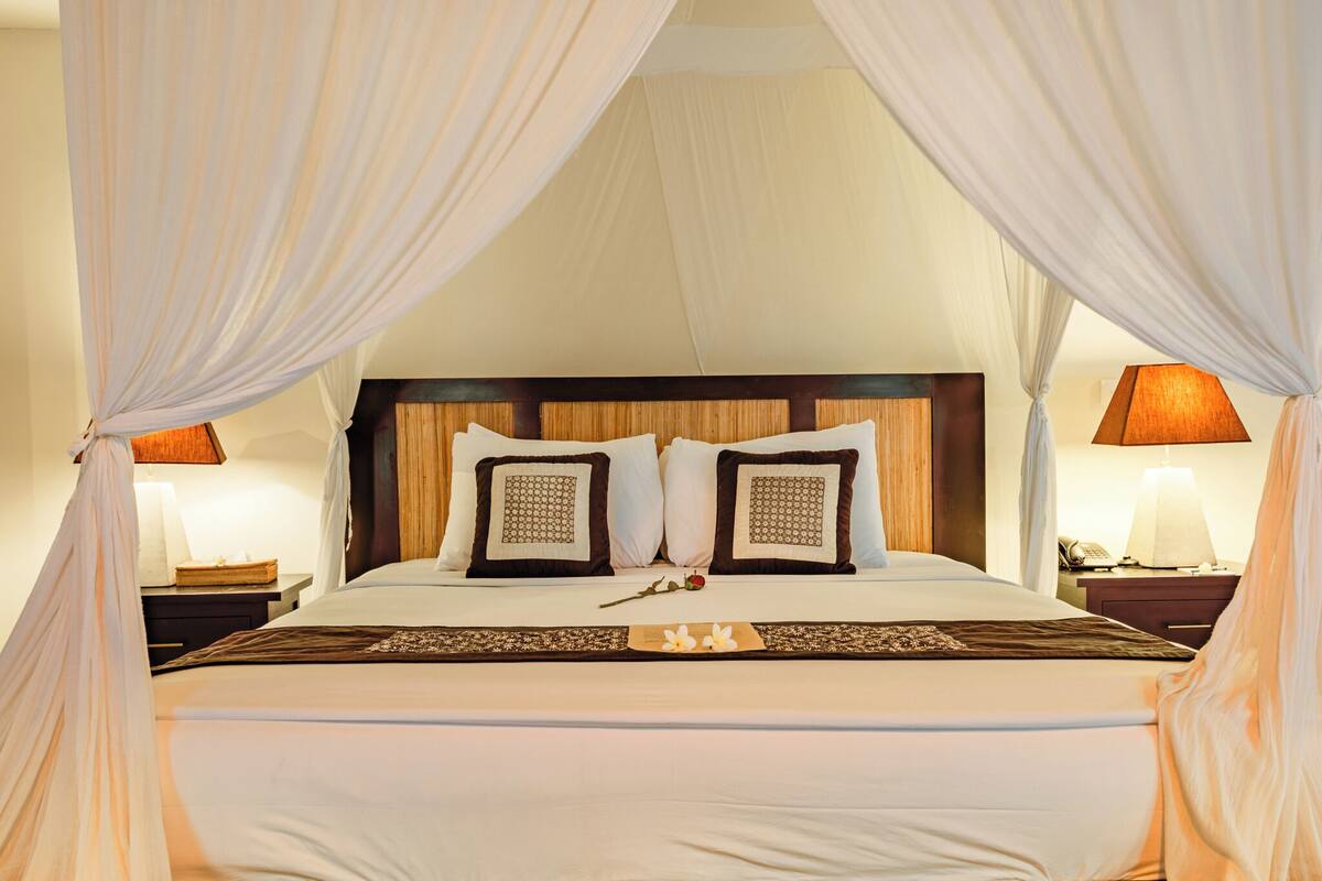 A neatly made bed with a wooden headboard, two decorative pillows, a bedside table with a lamp, and a canopy with draped fabric.