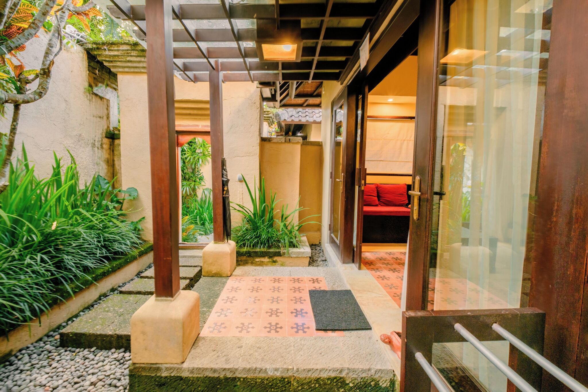 A narrow pathway leading to a room with a red sofa, surrounded by greenery and a tiled floor.