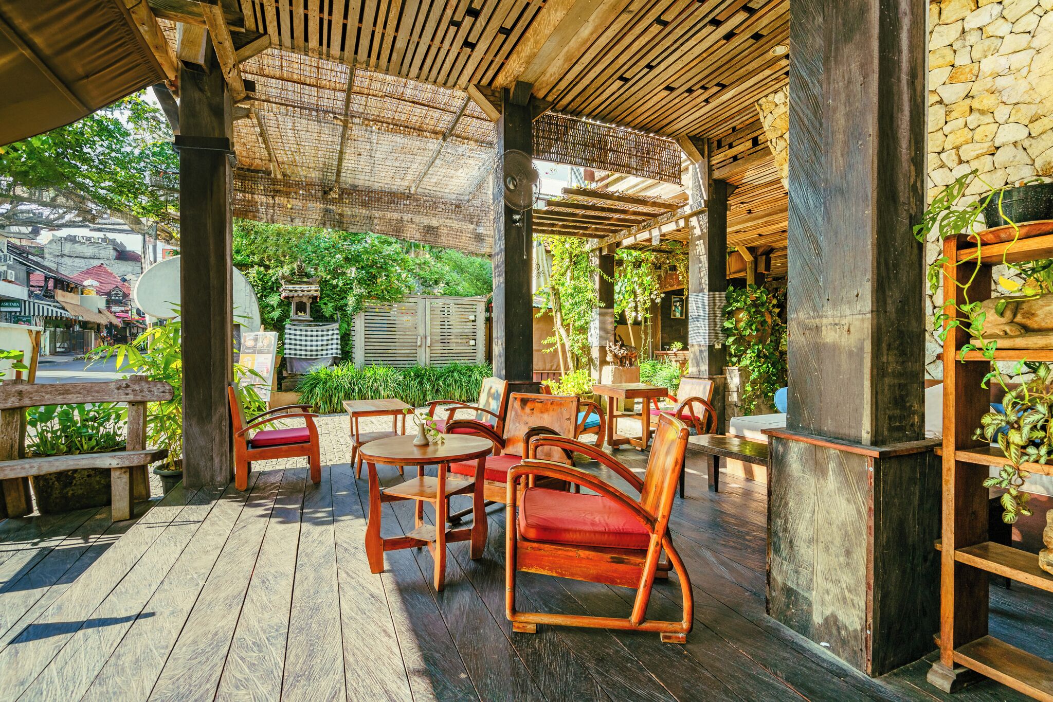 Una terraza exterior de madera con mesas y sillas, rodeada de vegetación y muros de piedra.