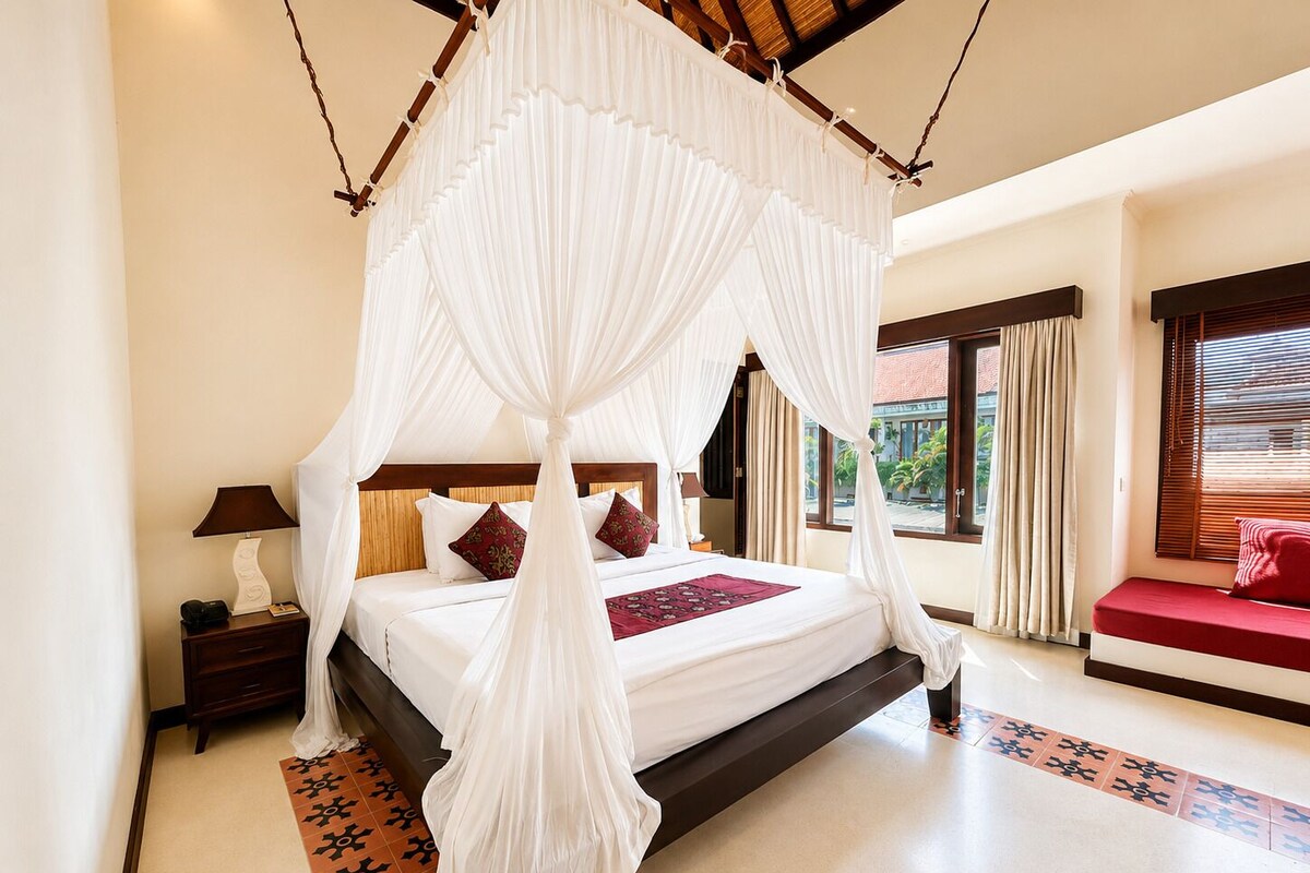 A bedroom with a four-poster canopy bed, a nightstand with a lamp, a bedside table, a red sofa, and a view of the outdoors through the windows.