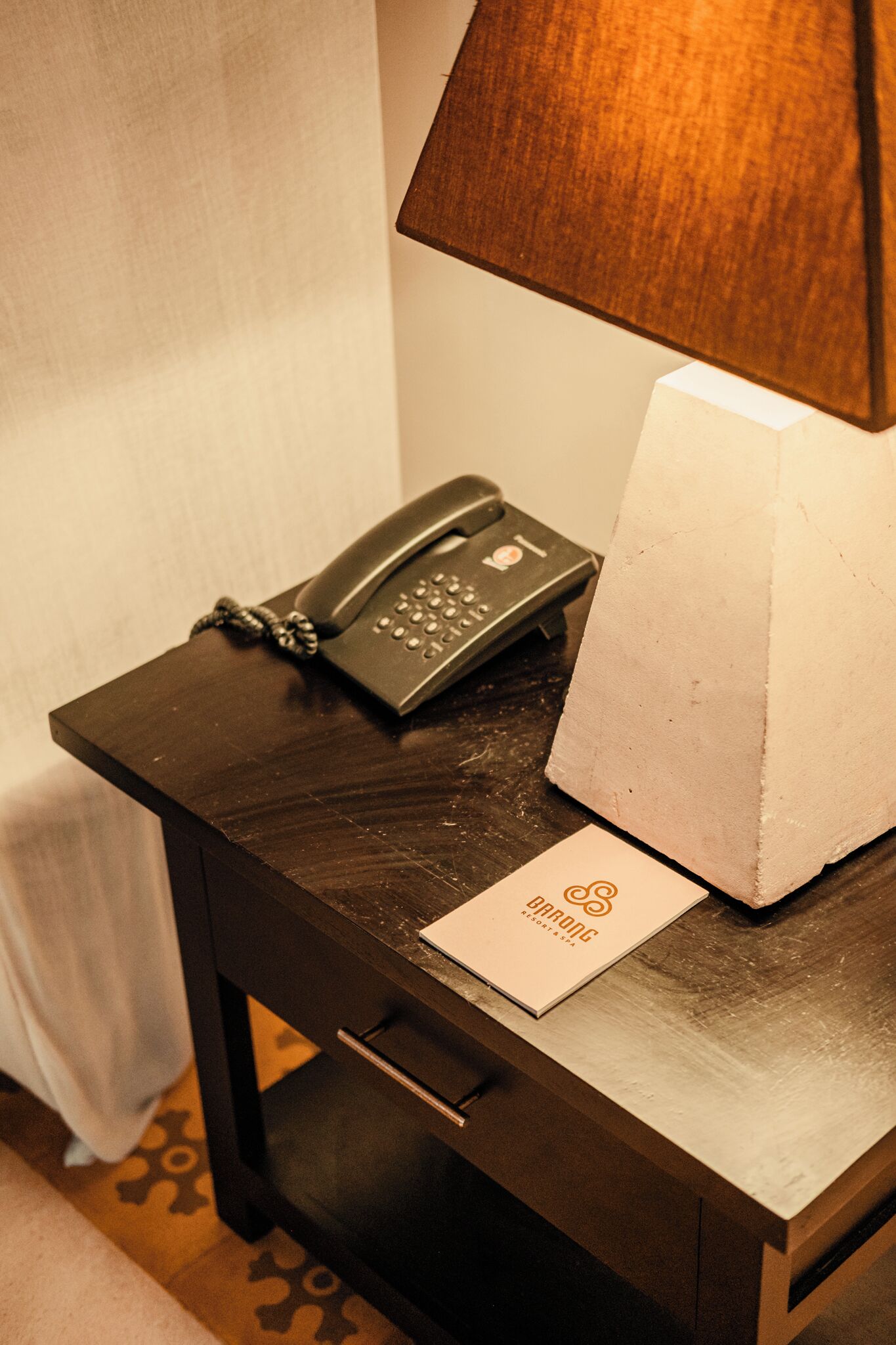 Una habitación de hotel con una mesita de noche de madera, un teléfono, una caja de pañuelos y un cartel con el nombre del hotel.