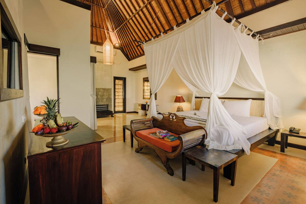 A bedroom with a four-poster bed, a wooden bench, a small table, and a fruit bowl.