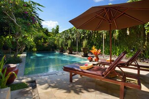 A poolside area with a lounge chair and an umbrella, surrounded by lush greenery.