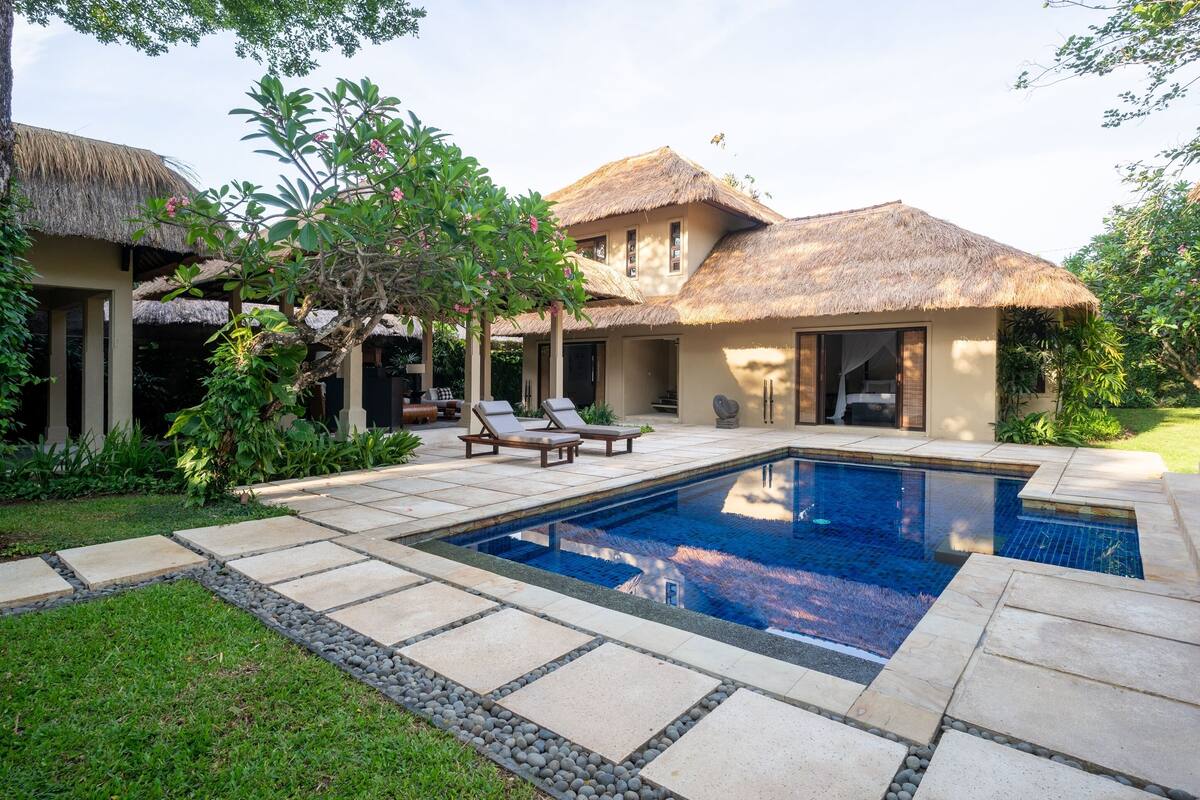 A pool area with lounge chairs, a thatched-roof building, and a well-maintained garden.