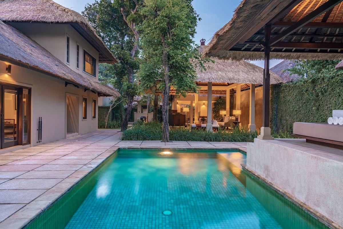 A swimming pool with a tiled edge, surrounded by a patio area with a thatched roof structure.