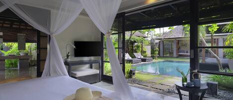 A bedroom with a bed, a TV, a small table, and a view of a swimming pool.