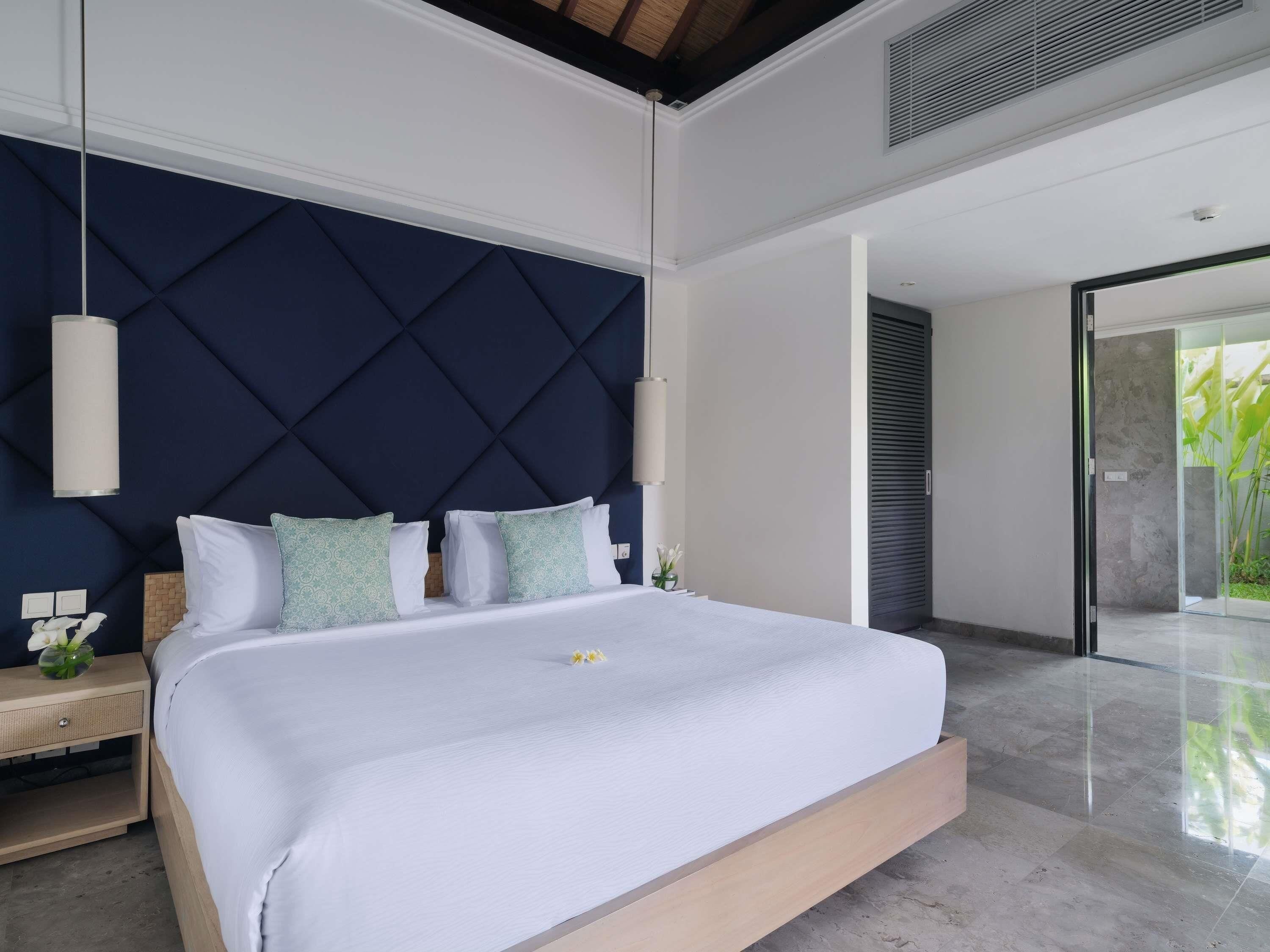 A modern bedroom with a large bed, a dark headboard, bedside tables, and a view of the outdoors.