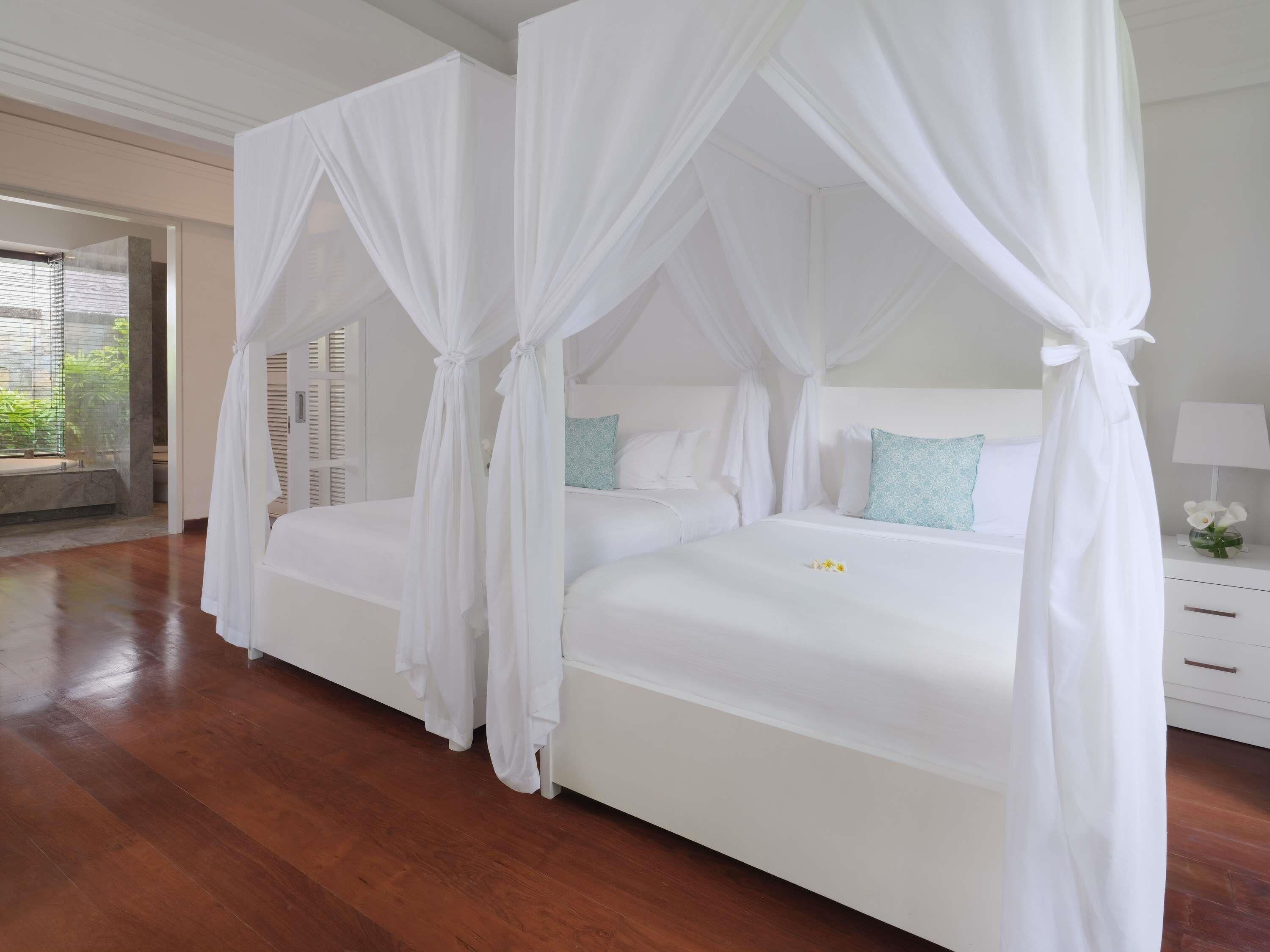 A bedroom with a four-poster canopy bed, wooden flooring, and a nightstand with a lamp and flowers.
