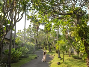 A pathway in a lush garden with a person in a long dress and hat walking away.