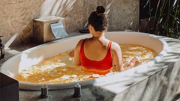 A person in a bathtub with a marble interior, surrounded by a stone wall and a plant.