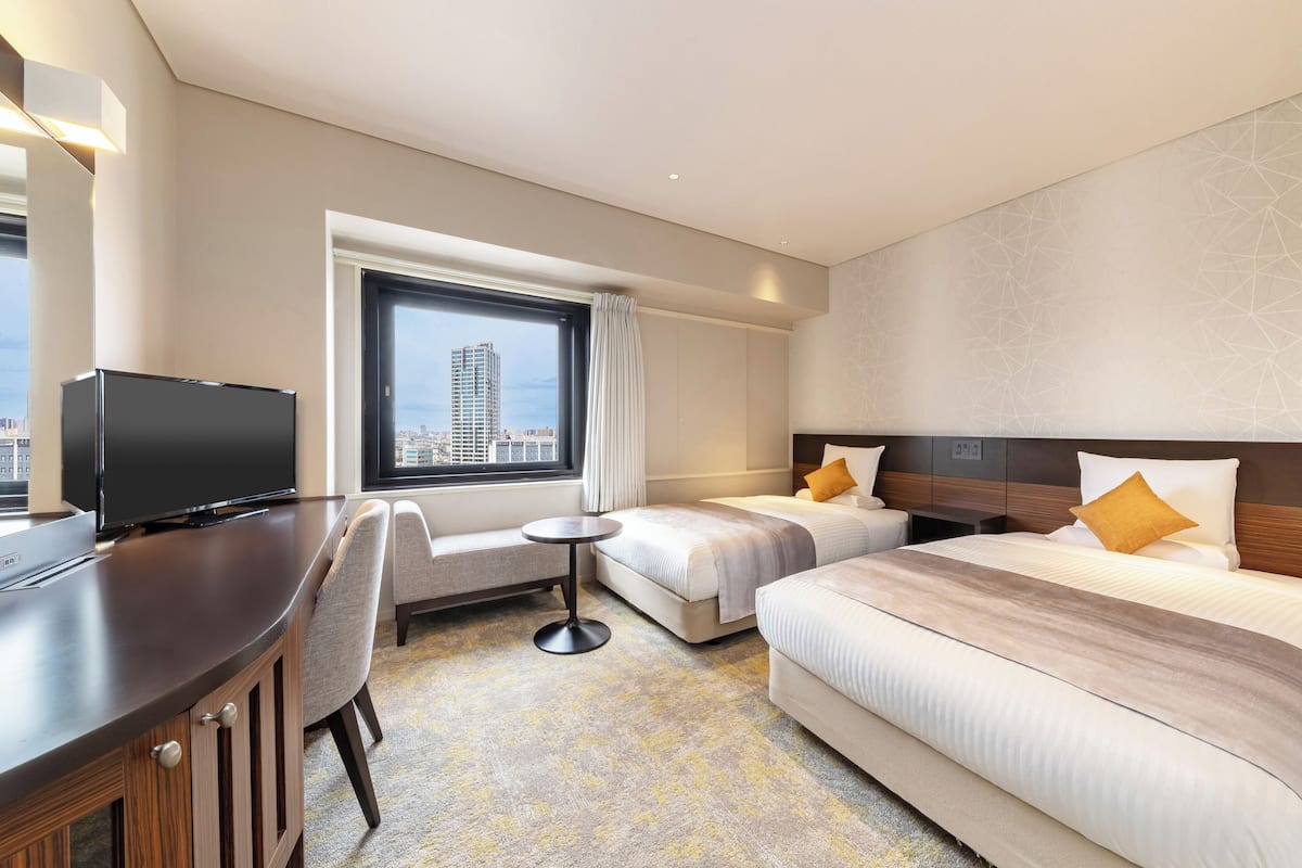 Ein Hotelzimmer mit Fernseher, zwei Betten, einem Schreibtisch und einem Fenster mit Blick auf die Stadt.