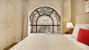 Habitación de hotel con una cama grande, una mesita de noche con lámpara y vistas a través de un ventanal decorativo.