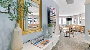 Una zona de recepción con una consola blanca, un espejo grande, una planta en maceta, un atril para revistas y un área de descanso con mesas y sillas.