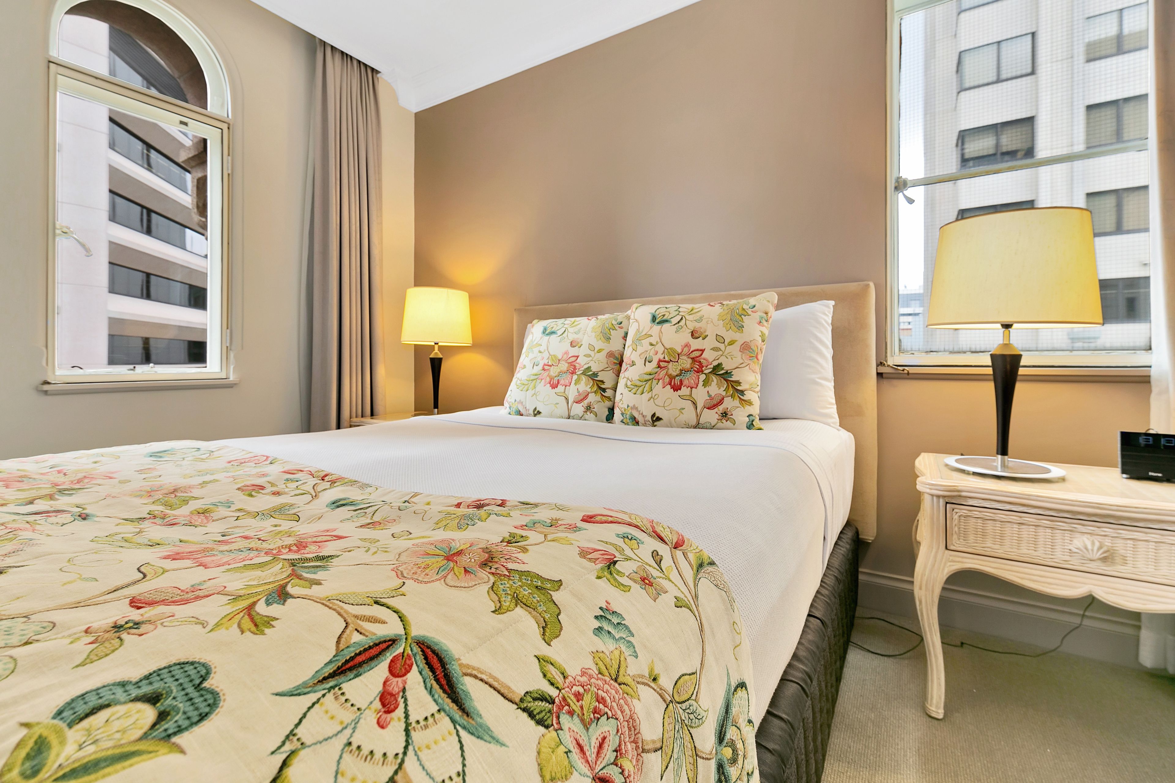 Hotelkamer met een bed, een bloemenprint beddeken, een nachtkastje met lamp, een raam met gordijnen en uitzicht op een gebouw.