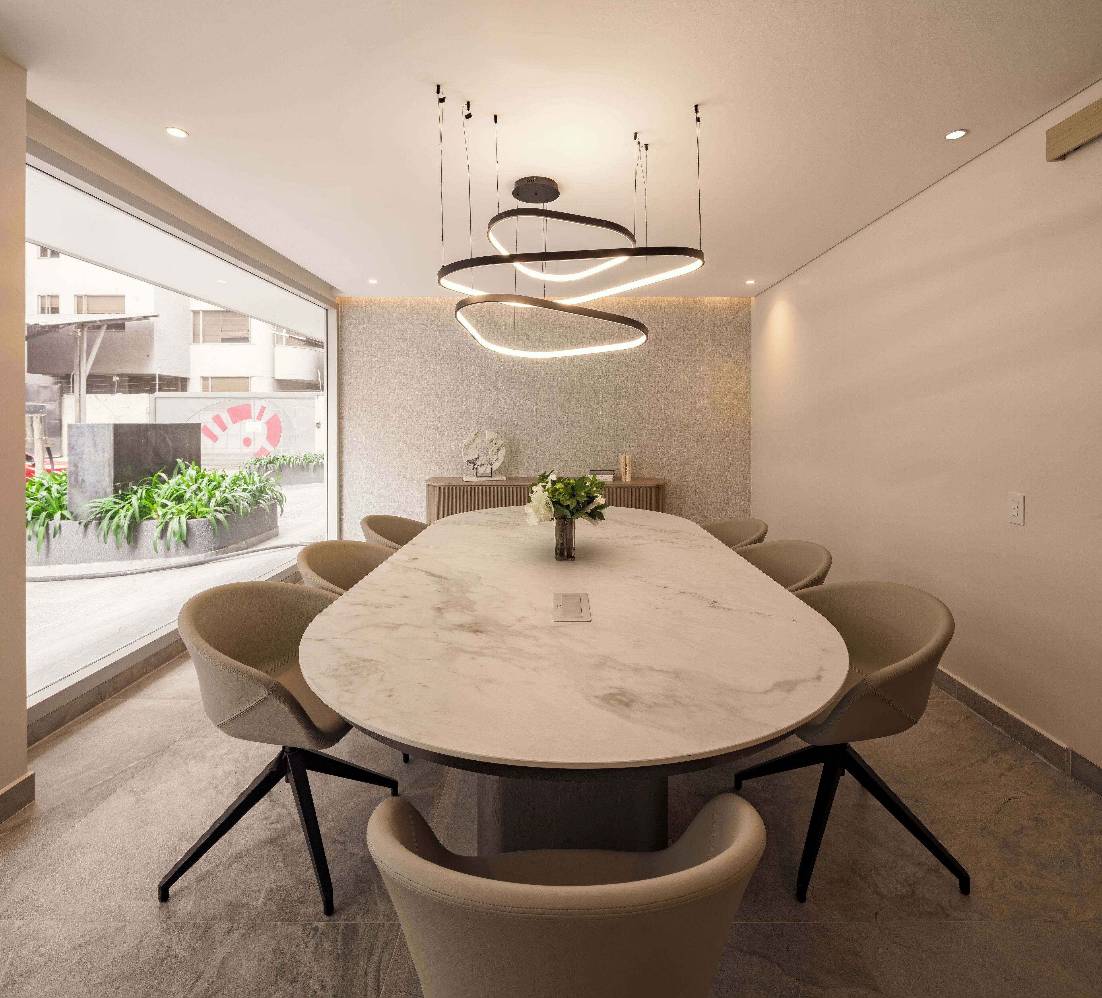 Une salle de conférence moderne, dotée d’une table en marbre, de chaises beiges, d’une plante au centre et d’un luminaire circulaire original au plafond.