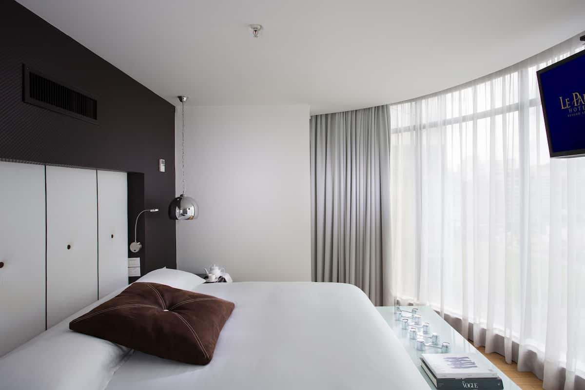Una habitación de hotel moderna con una cama grande, un televisor de pantalla plana, una mesita de noche con lámpara y vistas a la ciudad a través de cortinas traslúcidas.