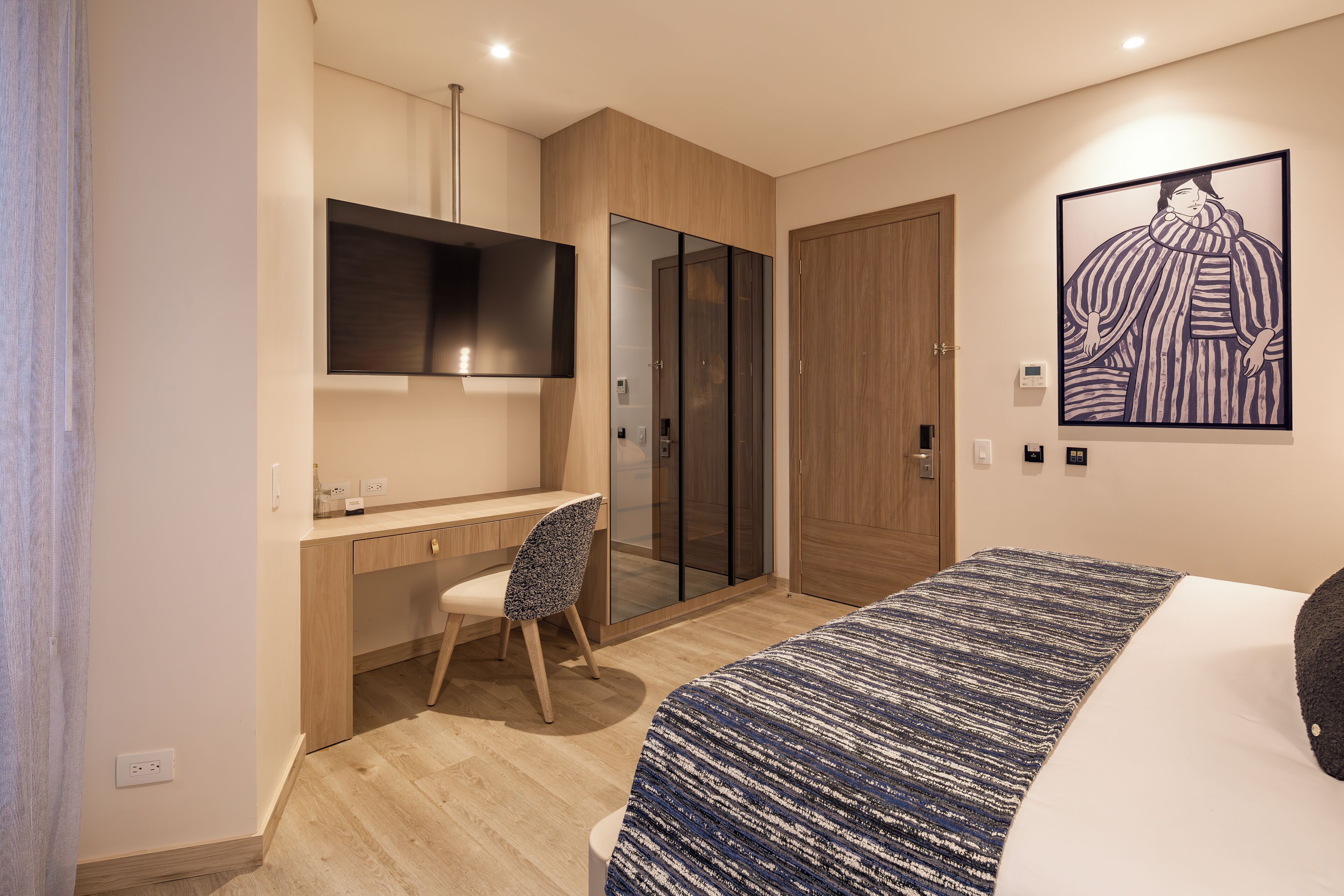 A modern hotel room with a wooden desk, a chair, a bed with a patterned bedspread, a large artwork on the wall, and a wardrobe with sliding doors.