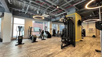 Un gimnasio con diversos equipos de ejercicio, amplias ventanas y un techo alto con tuberías a la vista.
