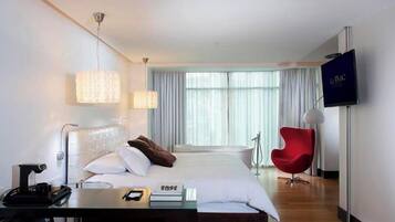 A hotel room with a bed, bedside table, lamp, TV, and a red chair.